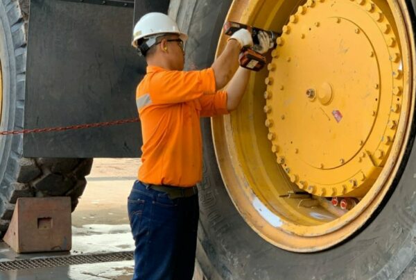 A worker in orange gear and a helmet tightens the bolts on the tire of a large construction vehicle.