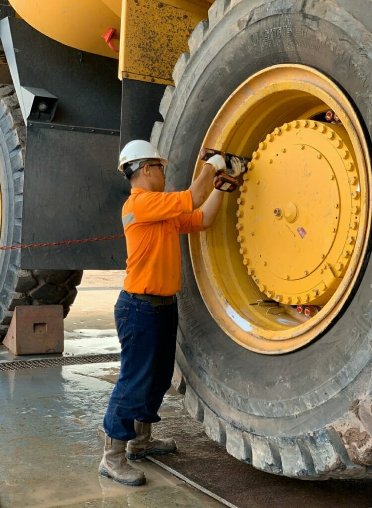 A worker in orange gear and a helmet tightens the bolts on the tire of a large construction vehicle.