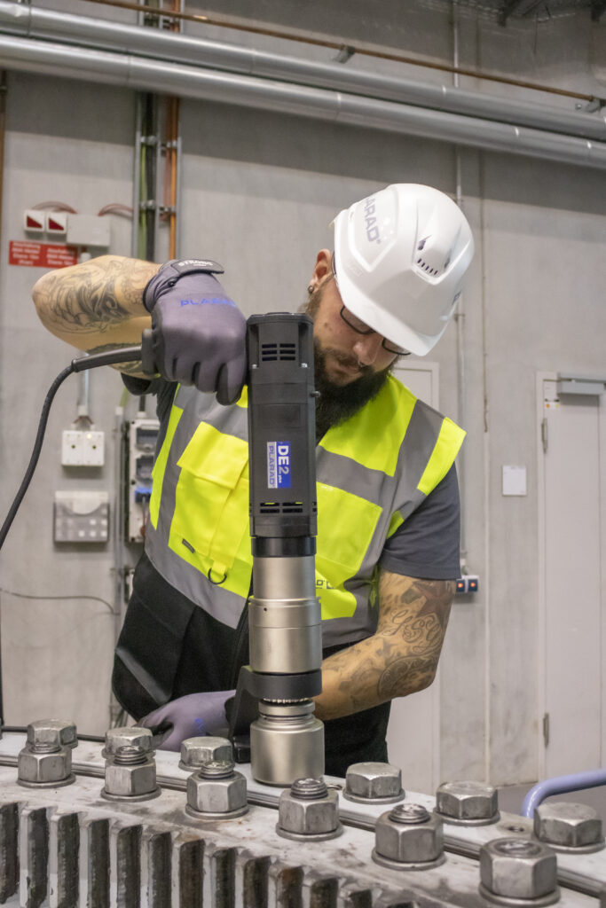 Ein Arbeiter mit Schutzhelm und Sicherheitsweste bedient fachmännisch den elektrischen Drehschrauber DE2power von Plarad und wendet dabei präzise Schraubtechnik an einem großen Industriegerät an.