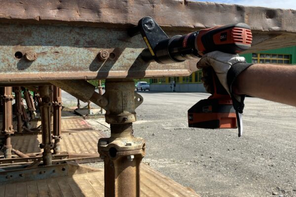 A person uses a DA2 battery nutrunner to repair or disassemble a rusted metal structure outdoors.