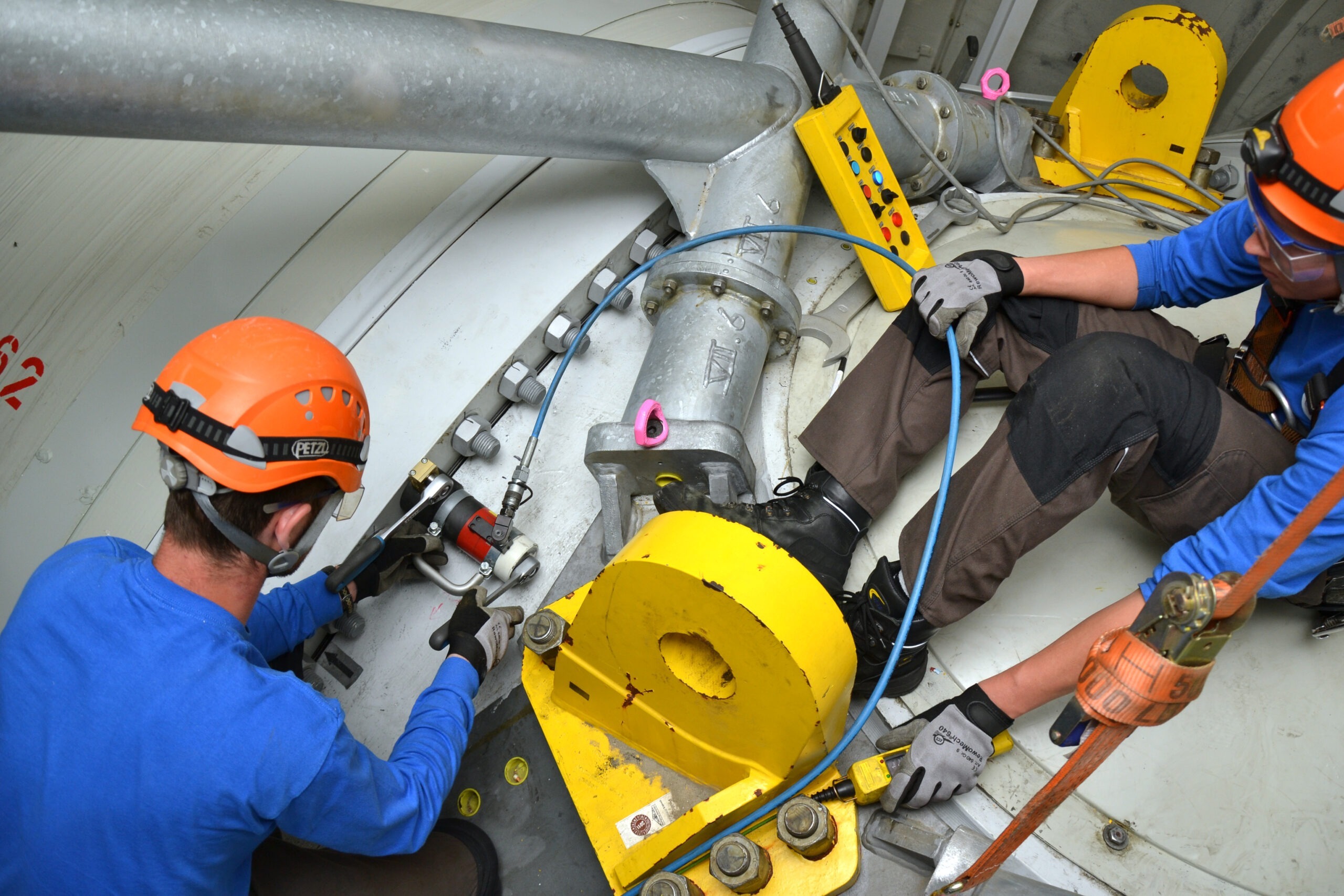 Two workers, wearing helmets and safety gear, carefully repair machinery in an industrial facility. They use precision bolting technology, hydraulic tensioners, and torque wrenches made in Germany to ensure optimal performance and safety.