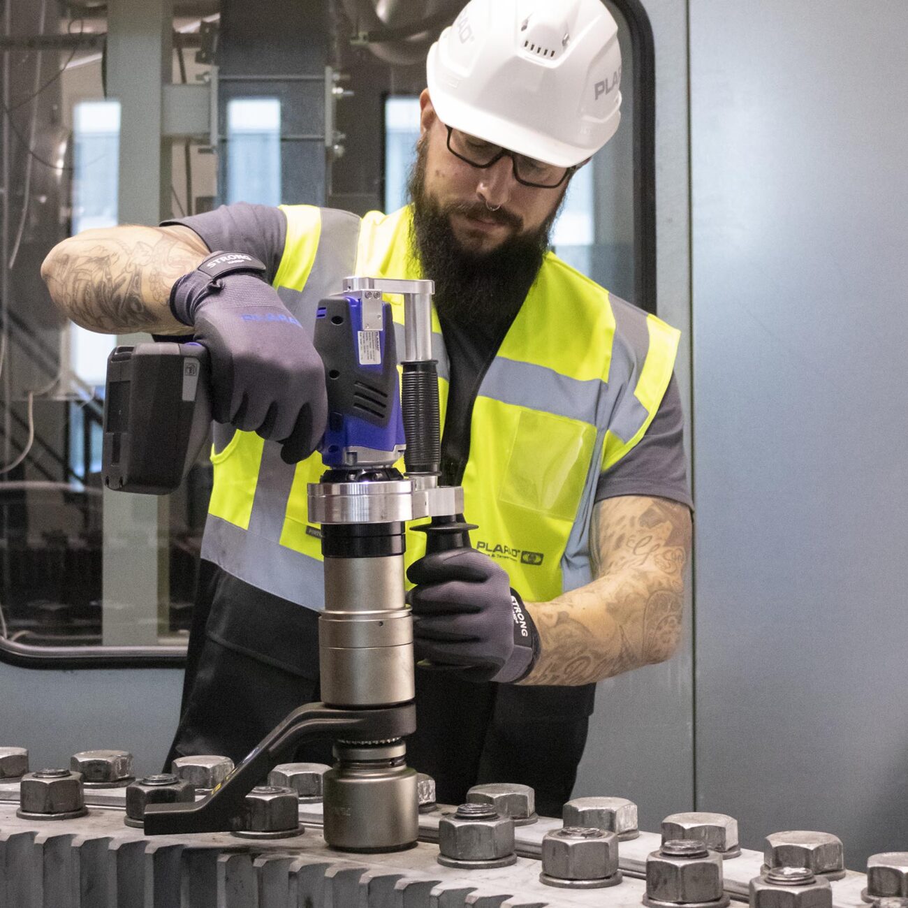 A worker wearing a hard hat expertly operates heavy machinery to tighten screws, using the innovative Plarad cordless nutrunner in an industrial setting. This safe fastening technology ensures precision and efficiency and underscores the importance of advanced technology in modern workplaces. 