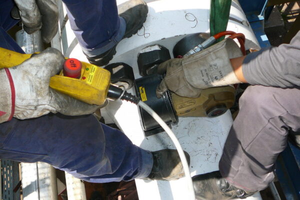 P1040295 Zwei Arbeiter der Öl, Gas & Chemischen Industrie verwenden einen hydraulischen Drehmomentschlüssel an Industrieanlagen. Sie tragen Handschuhe und handhaben geschickt ein Steuergerät.