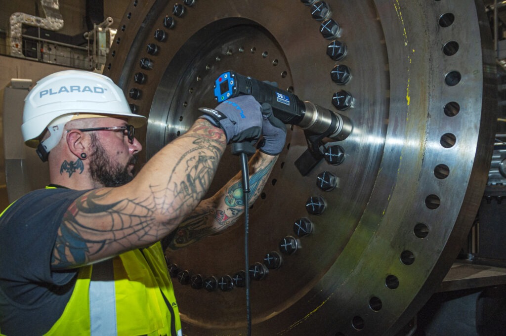 A man wearing a construction helmet precisely operates the Plarad DE1power electric nutrunner on a large industrial machine, surrounded by bolts.