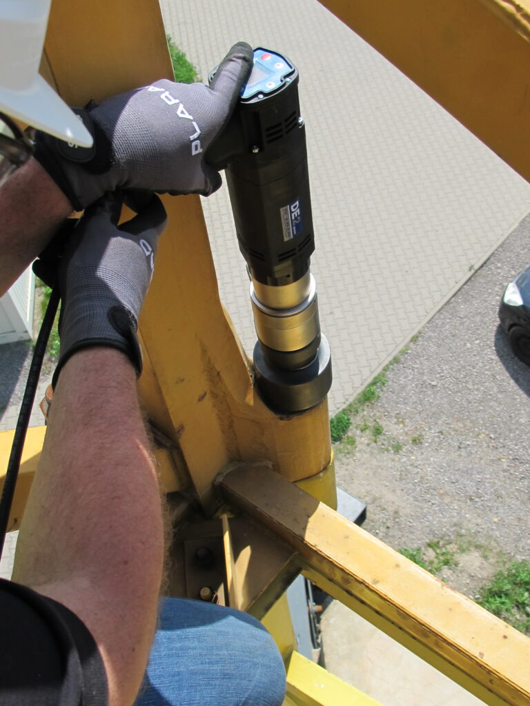 The person skillfully operates machinery on a metal structure, wearing gloves and a helmet. They precisely use the Plarad DE2power electric nutrunner while positioned above a paved surface below.