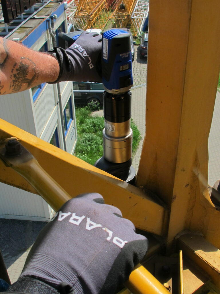 A person wearing gloves expertly operates the Plarad DE2power electric nutrunner, on the bolts of a crane structure.