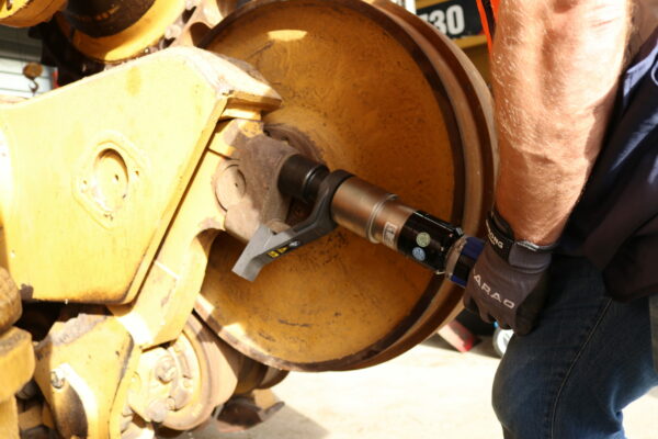 A person tightens a bolt on a heavy machine with large yellow wheels using a battery-powered torque wrench and screw technology from PLARAD