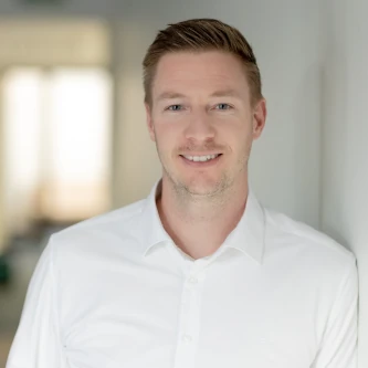 A smiling man in a white shirt stands confidently in a well-lit hallway, embodying Plarads commitment to around-the-clock service through its global distribution network.