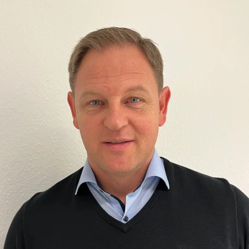 Man in a blue collared shirt and dark sweater, embodying the professionalism of Plarads service rund um die Uhr durch weltweites Vertriebsnetz, stands confidently against a plain white background.