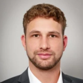 A man with short curly brown hair and a beard, wearing a suit and tie, stands ready to connect against a gray background, embodying Plarad Kontakte für Deutschlands commitment to being available rund um die Uhr.