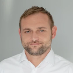 A man with short hair and a beard, wearing a white shirt, looks towards the camera with a neutral expression, embodying the reliability of Plarad Kontakte für Deutschland rund um die Uhr.